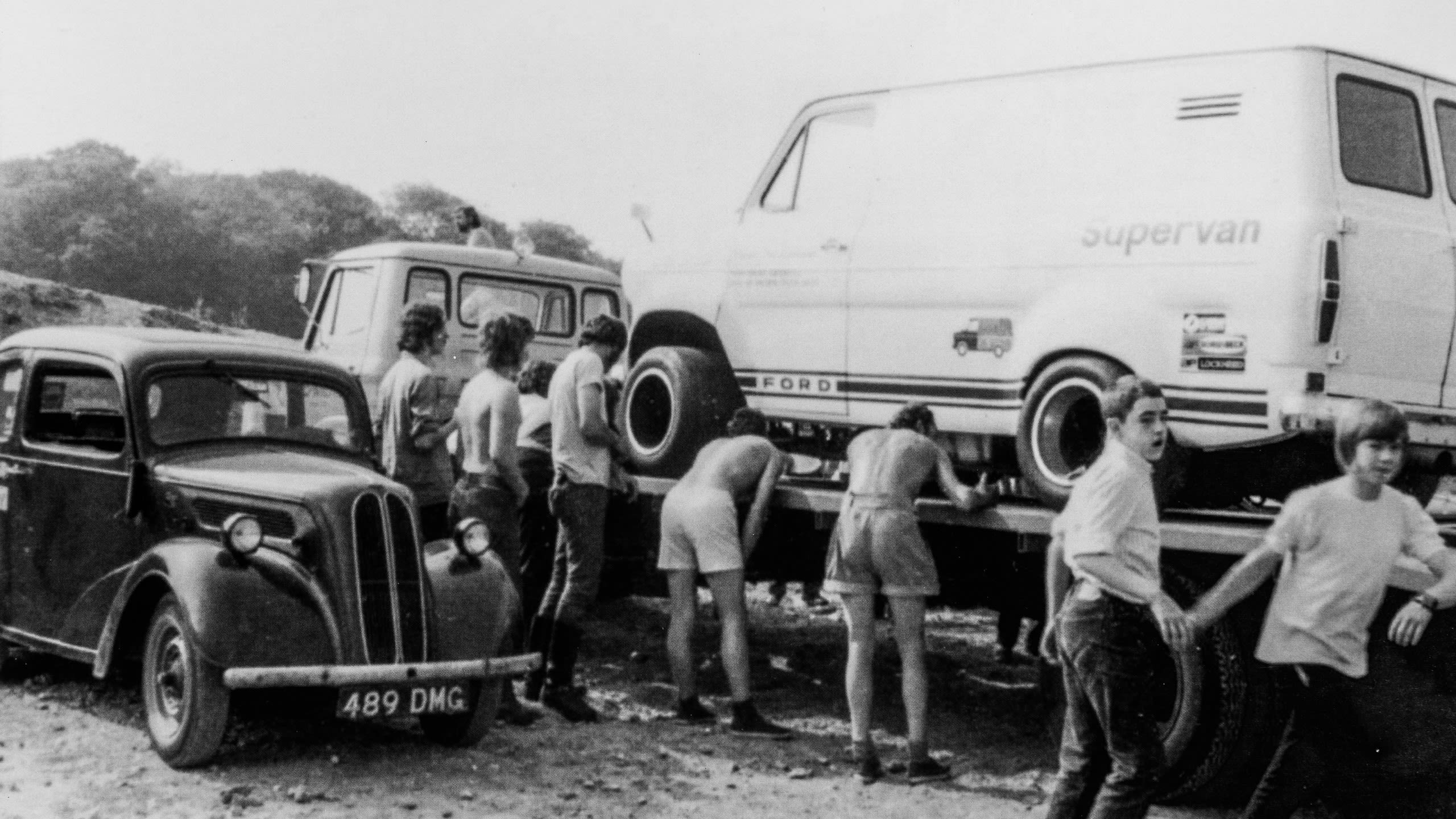 Archive image of a group of friends loading Supervan 1 onto the back of a flatbed truck. Teenagers look on next to a Ford Popular classic car from the 1950s.