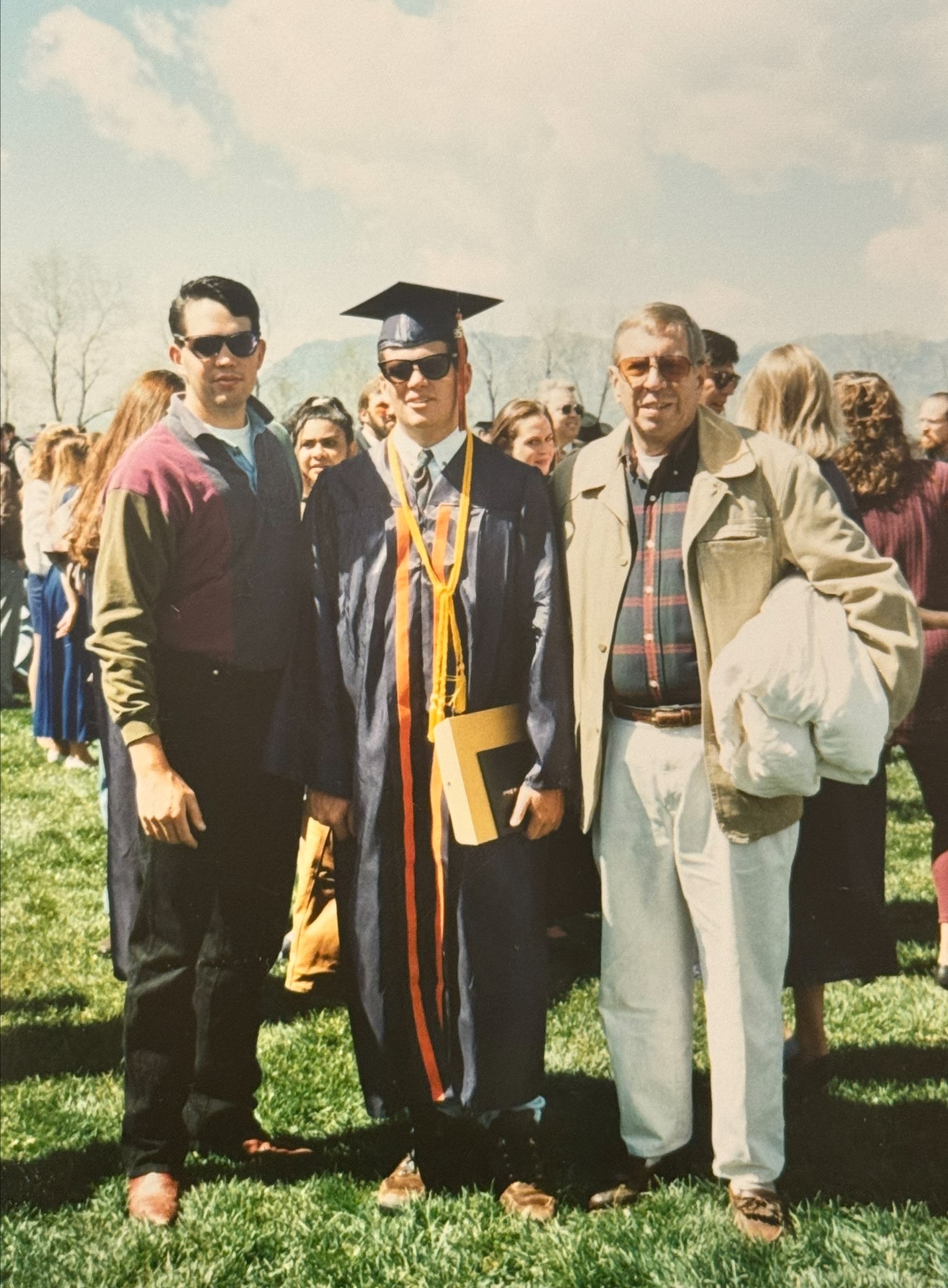 Two young men pose with an older man in an old photograph. One of the young men is wearing a graduation cap and gown. 