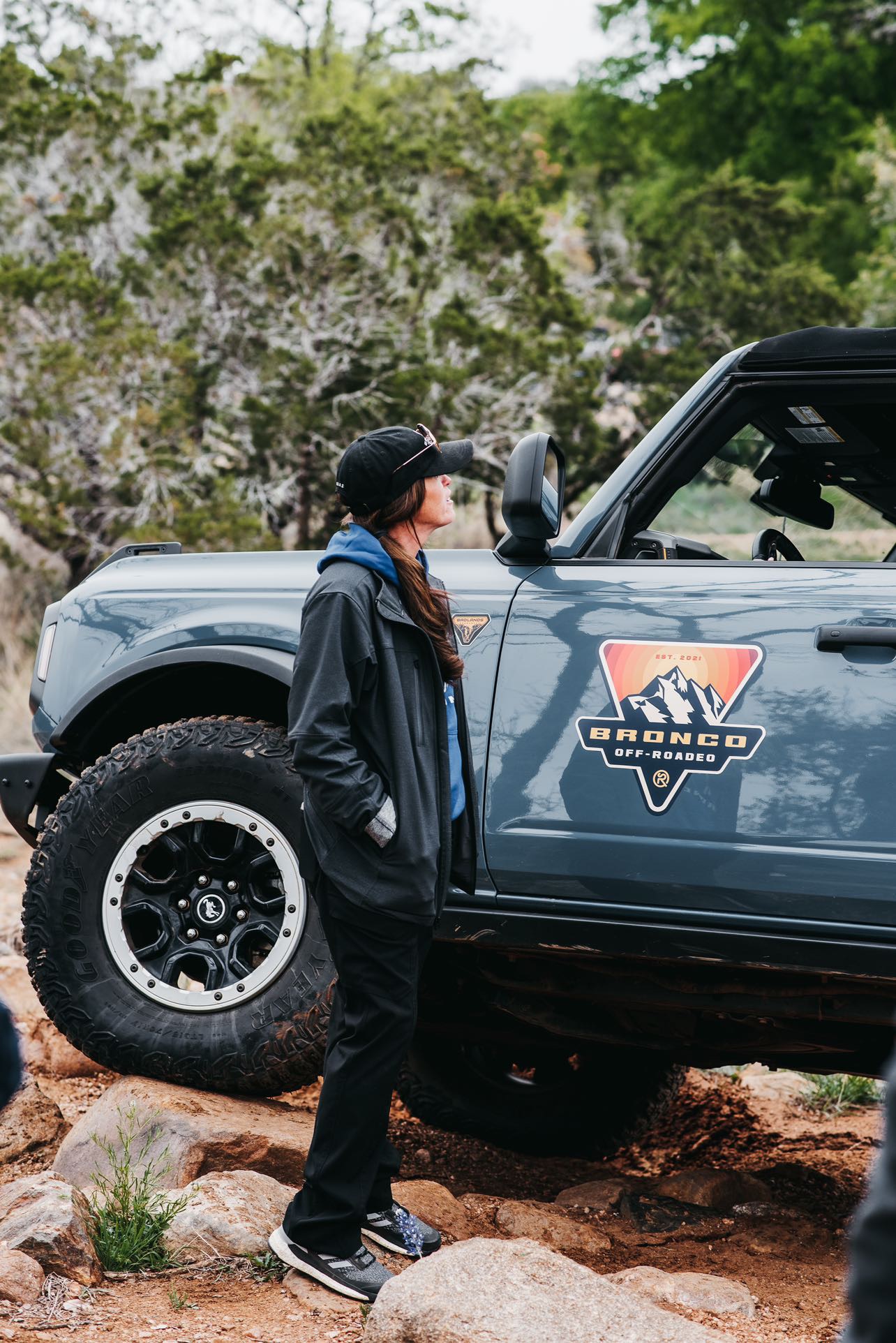 A woman stands next to a Bronco with a "Bronco Off-Roadeo" decal on the side. The Bronco is on rocky terrain and there is a forested background.