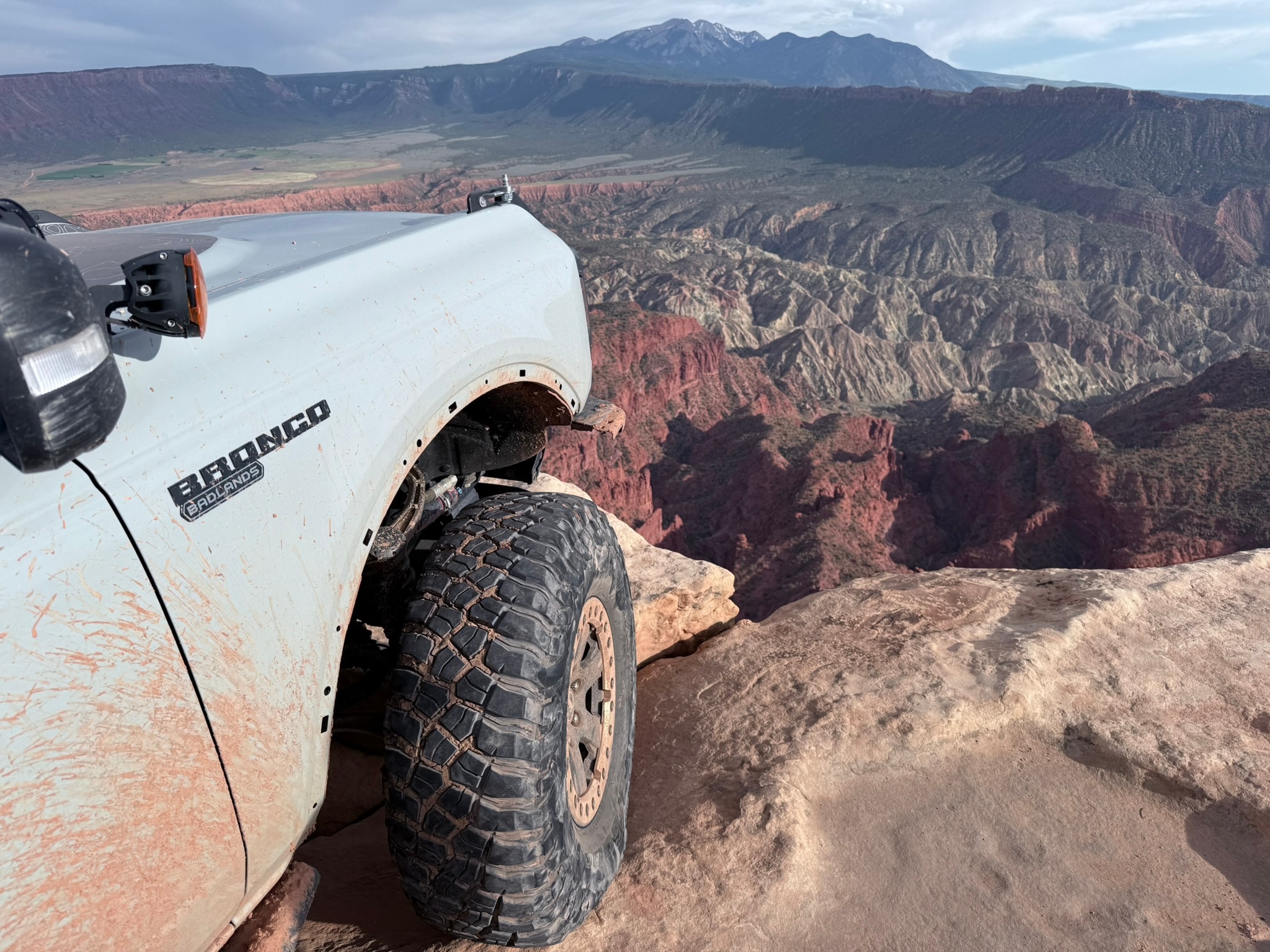 A close-up view of the front side and wheel of a Bronco parked at the edge of a cliff. Red rock can be seen far below.