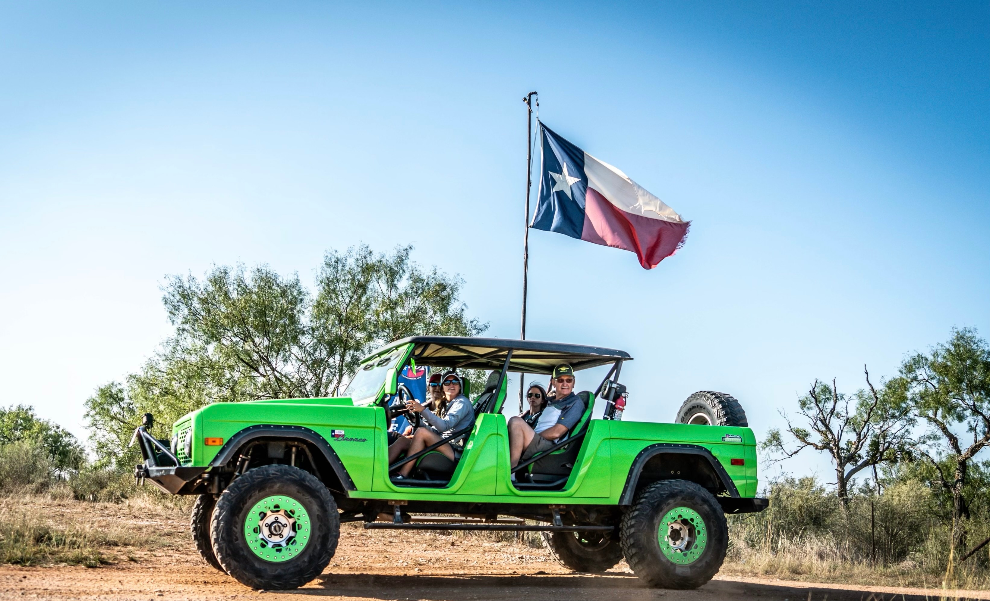 A group of people look at the camera from inside a lime green Bronco with a Texas flag waving on top.