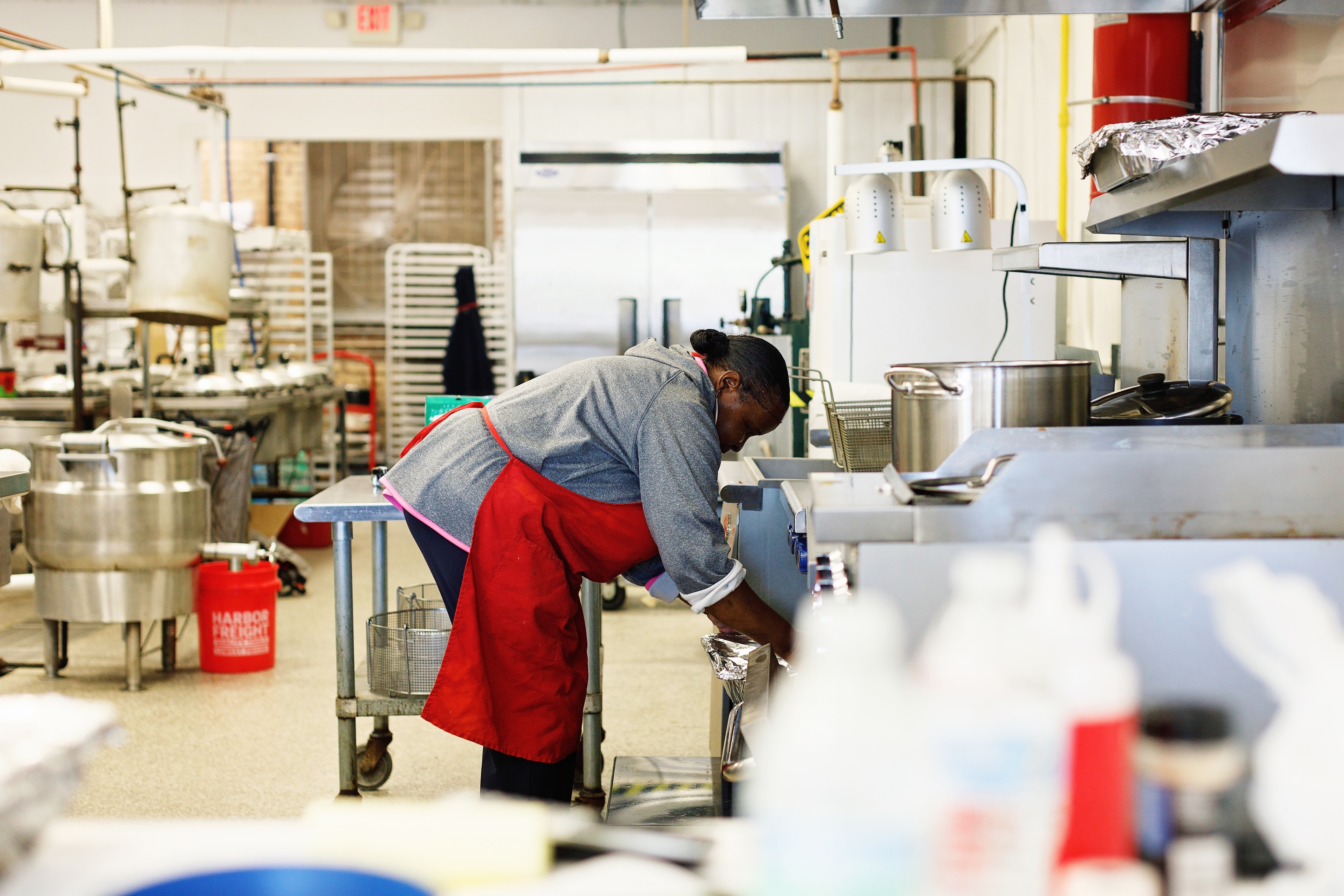 A woman wearing a red apron leans over a countertop in a commercial kitchen.