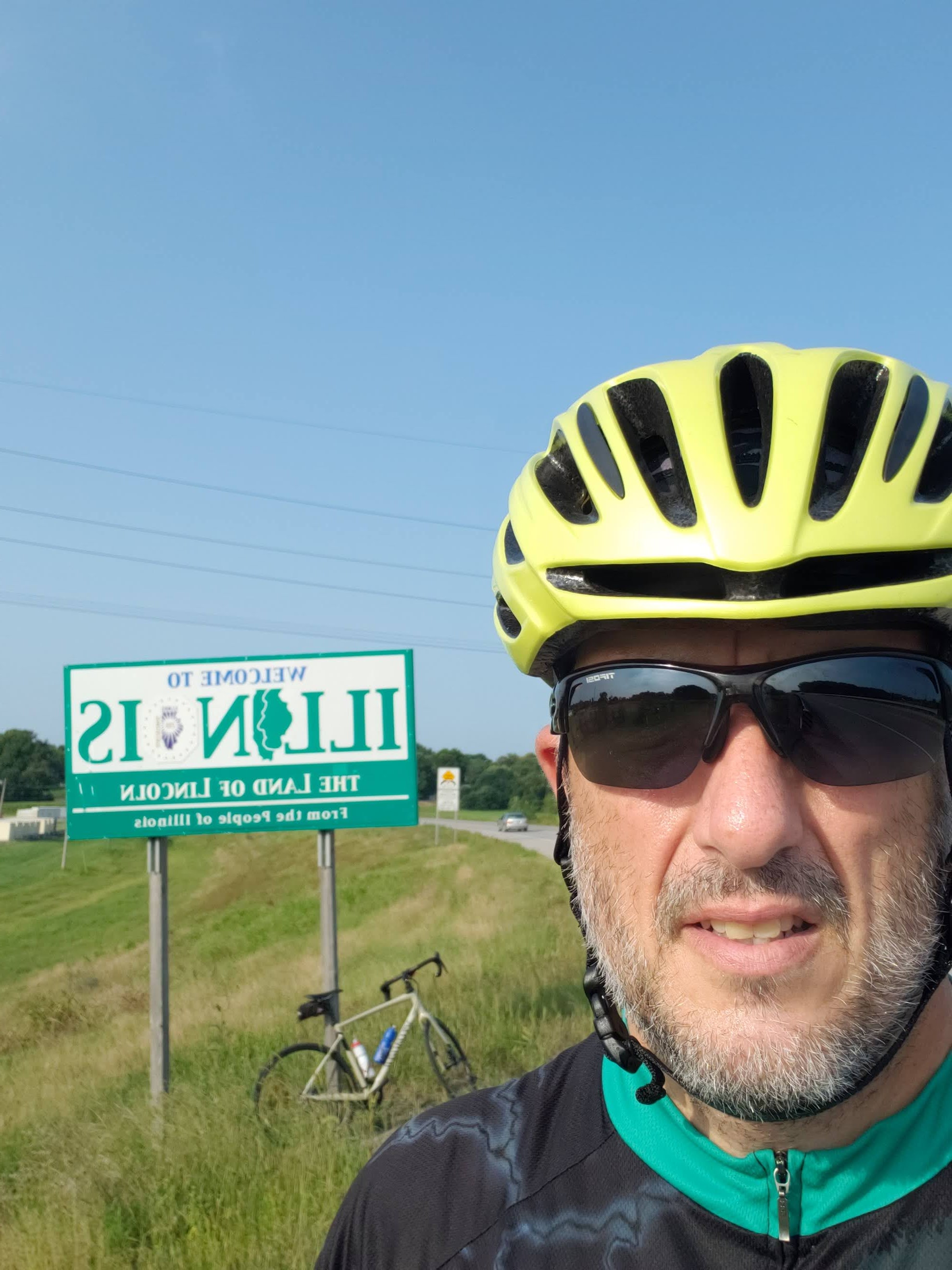 A man in a bicycle helmet takes a selfie in front of the Illinois state sign.
