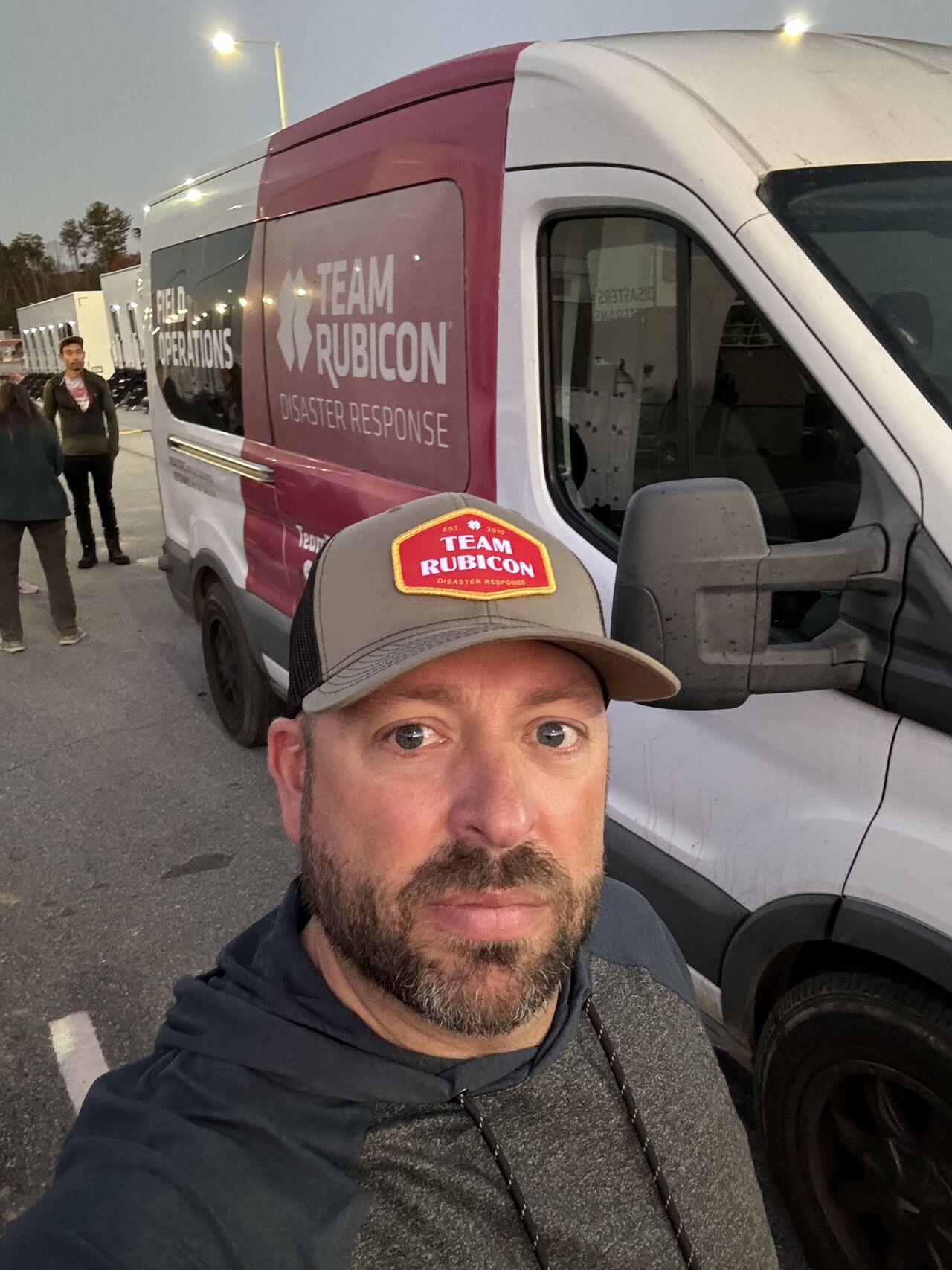 A man wearing a baseball cap that reads "Team Rubicon" stands in front of a van with "Team Rubicon: Disaster Response" written on the side.