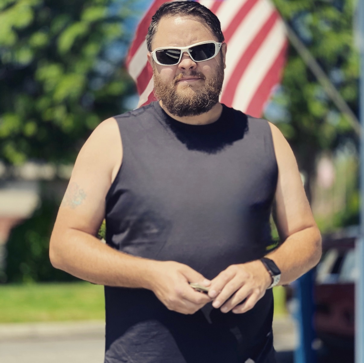 A man in sunglasses and a muscle tee poses in front of an American flag.