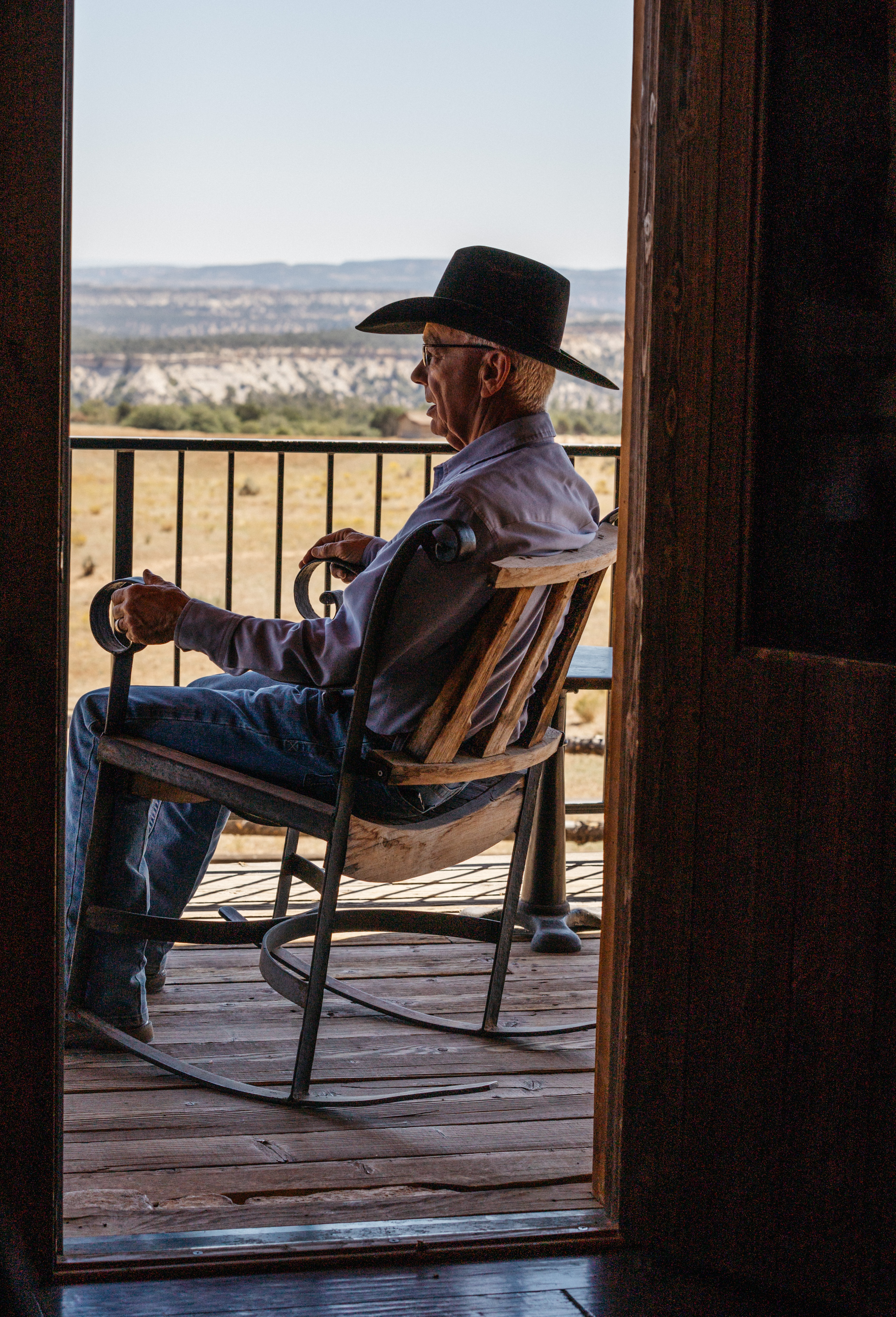 A man in a cowboy hat sits in a rocking chair on the porch. The view is from behind, and in front of him you can see a vast expanse of land.