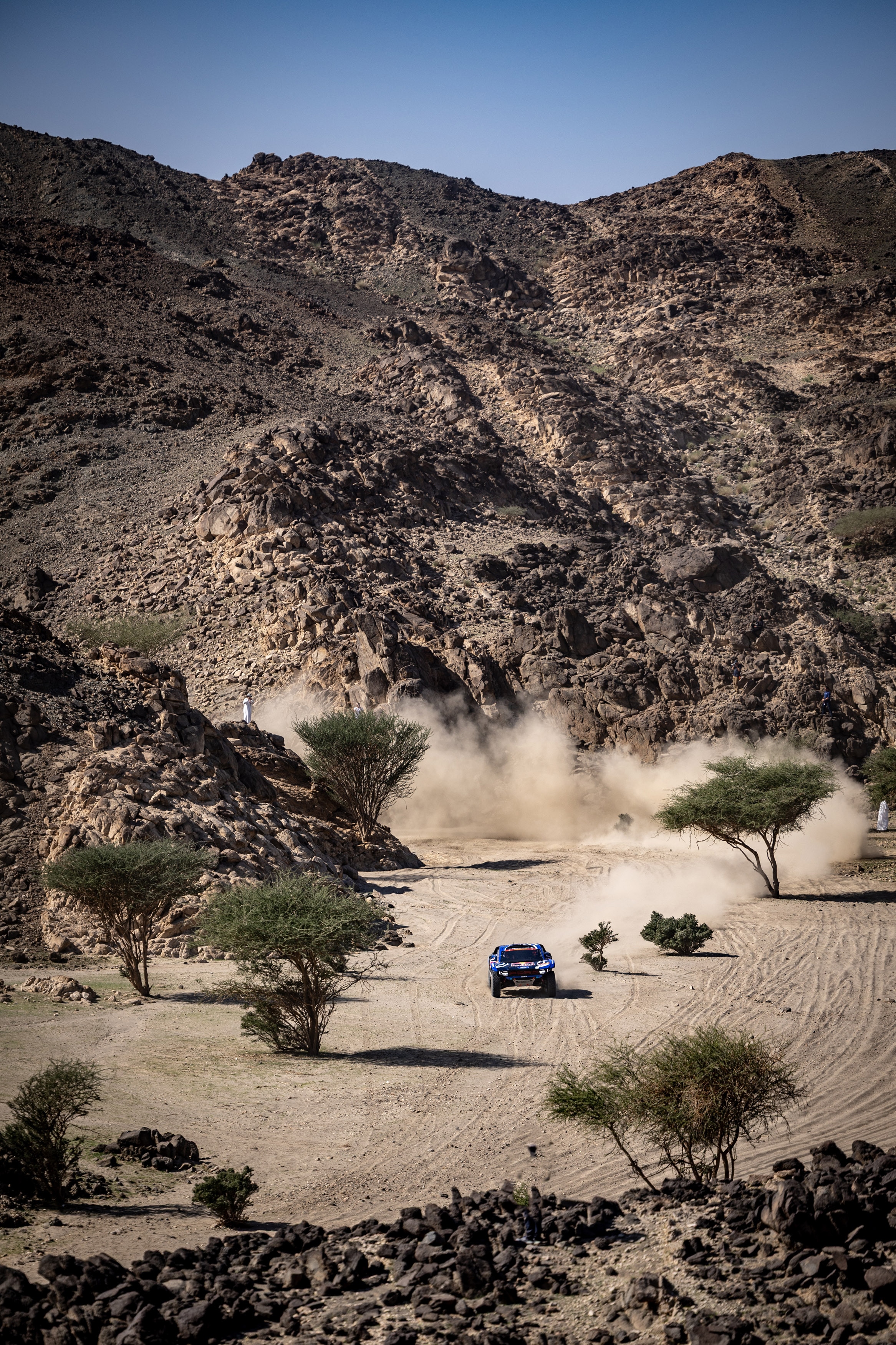 A Ford off-road vehicle drives through the desert. The backdrop of the photo includes a blue sky over a scenic ridge.